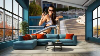 Pretty joyful woman with long black hair standing by a metal fence of a swimming pool wearing white swimwear and sunglasses. concept of luxury holidays Wall mural