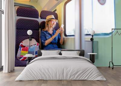 Enjoying travel. Young pretty woman traveling by the train sitting near the window using smartphone. Wall mural