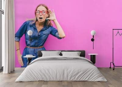 Gossip concept. Colorful portrait of amazed young woman with palm near her ear. Pink background. Wall mural