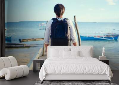 Photography and travel. Young man with rucksack holding camera standing on wooden fishing pier enjoying beautiful tropical sea view. Wall mural