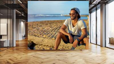 Relaxed and cheerful. Outdoor portrait of happy young african man sitting near the sea. Wall mural