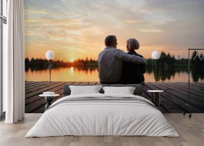 Romantic holiday. Senior loving couple sitting together on lake bank enjoying beautiful sunset. Wall mural