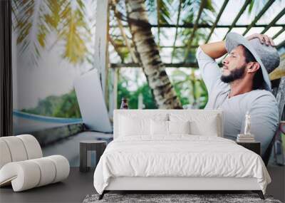 Technology and travel. Working outdoors. Freelance concept. Bearded young man using laptop in cafe on tropical beach. Wall mural