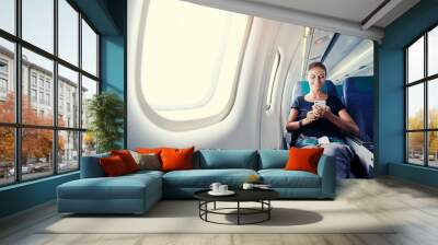 Travel and technology. Young woman in plane using smartphone while sitting in airplane seat. Wall mural