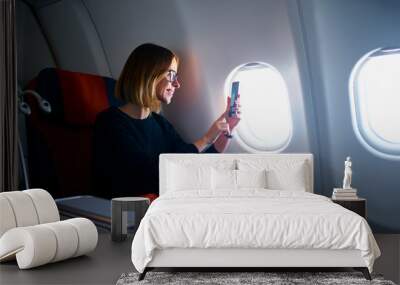 Traveling and technology. Flying at first class. Pretty young businees woman taking photo on smartphone while sitting in airplane. Wall mural