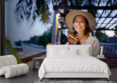 Vacation and technology. Young pretty woman in hat using smartphone sitting at beach cafe bar. Wall mural