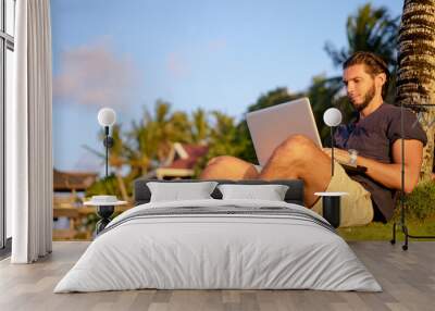 Work and vacation. Young man working on laptop computer on the tropical beach under the palm tree. Wall mural