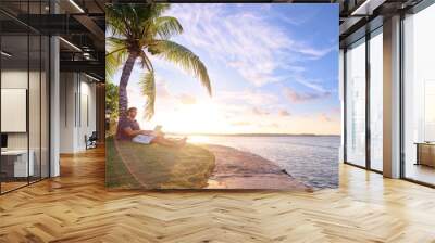 Work and vacation. Young man working on laptop computer on the tropical beach under the palm tree. Wall mural