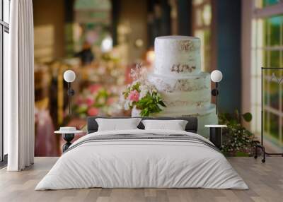 a beautiful table adorned with small flower plants surrounding a delicious white three-tier cake which also has flowers in the middle, in the background are more arrangements and large windows in a ha Wall mural