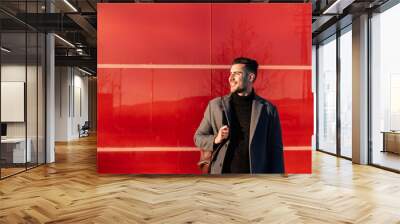 Young businessman with backpack smiling in front of a red wall at sunset Wall mural