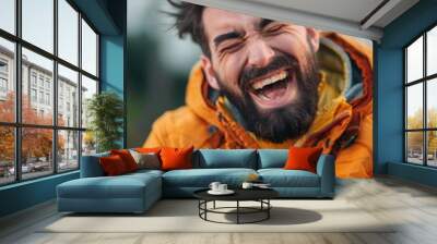 Man with beard laughing while looking at cellphone. Outdoor setting, possibly park or garden. Casual attire with hooded jacket. Wall mural