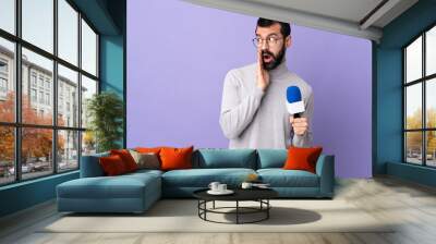 Adult reporter man with beard holding a microphone over isolated purple background whispering something with surprise gesture while looking to the side Wall mural