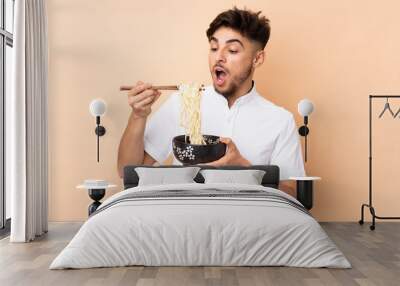 Arabian handsome man isolated on beige background holding a bowl of noodles with chopsticks and eating it Wall mural