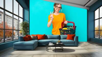 Blonde English young girl holding a shopping basket full of food isolated on blue background happy and counting three with fingers Wall mural