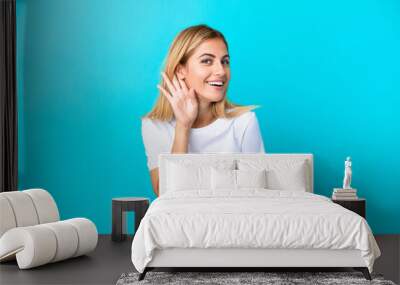 Blonde Uruguayan girl isolated on blue background listening to something by putting hand on the ear Wall mural