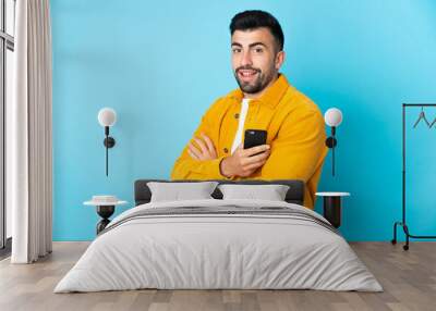 Caucasian man over isolated blue background holding a mobile phone and with arms crossed Wall mural