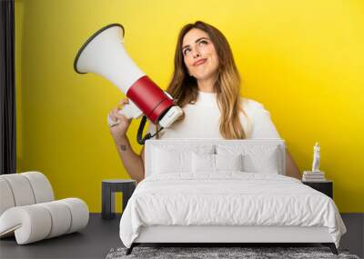 Caucasian woman isolated on yellow background holding a megaphone and thinking Wall mural