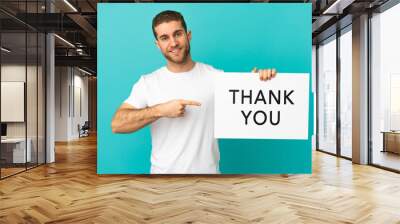 Handsome blonde man over isolated blue background holding a placard with text THANK YOU and  pointing it Wall mural