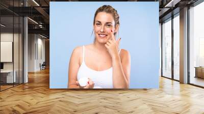 Happy Young blonde woman with moisturizer over isolated blue wall Wall mural