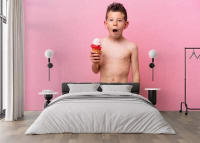 Little caucasian boy eating an ice-cream isolated on pink background with surprise and shocked facial expression Wall mural