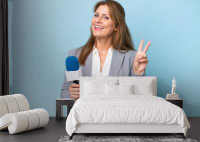 Middle-aged TV presenter woman over isolated blue background smiling and showing victory sign Wall mural