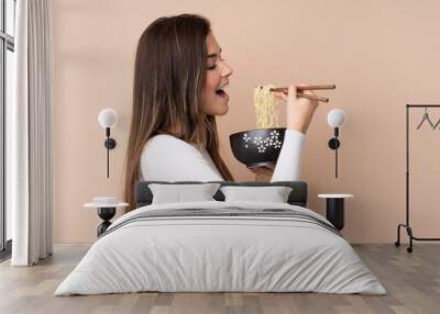 Teenager girl over isolated background holding a bowl of noodles with chopsticks and eating it Wall mural