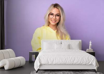 Teenager girl wearing a yellow sweatshirt over isolated purple background with glasses and smiling Wall mural