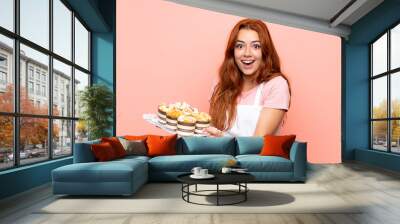 Teenager redhead girl holding lots of different mini cakes over isolated pink background Wall mural