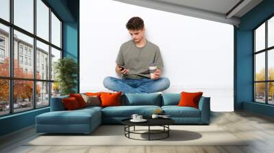 Teenager Russian man sitting on the floor isolated on white background holding coffee to take away and a mobile Wall mural