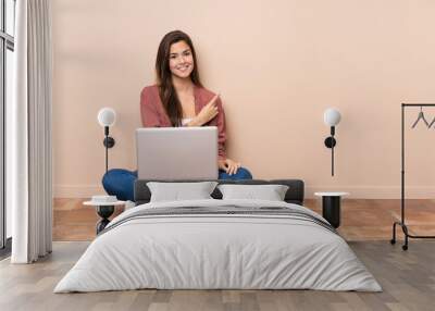 Teenager student girl sitting on the floor with a laptop pointing to the side to present a product Wall mural