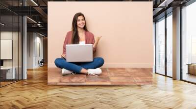 Teenager student girl sitting on the floor with a laptop pointing to the side to present a product Wall mural