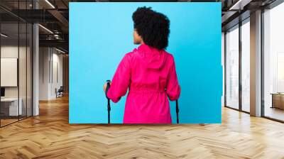 Young Africa American with backpack and trekking poles isolated on blue background in back position and looking side Wall mural