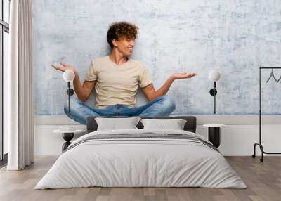 Young african american man sitting on the floor holding copyspace with two hands Wall mural