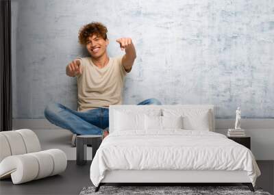 Young african american man sitting on the floor points finger at you while smiling Wall mural