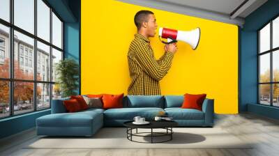 Young afro american man on yellow background shouting through a megaphone to announce something in lateral position Wall mural