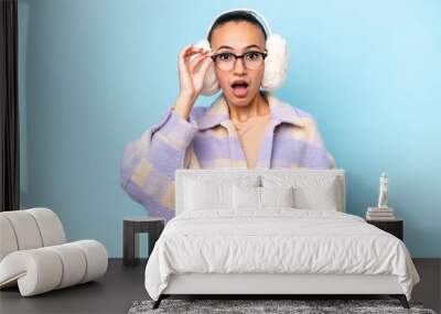 Young Arab woman wearing winter muffs isolated on blue background with glasses and surprised Wall mural