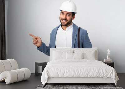 Young architect man with helmet and holding blueprints isolated on white background pointing finger to the side Wall mural