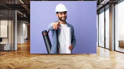 Young architect Moroccan man with helmet and holding blueprints over isolated background pointing front with happy expression Wall mural