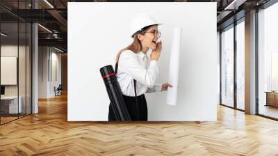 Young architect woman with helmet and holding blueprints isolated on white background shouting with mouth wide open to the side Wall mural