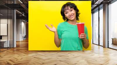 Young Argentinian woman holding a soda isolated on yellow background extending hands to the side for inviting to come Wall mural