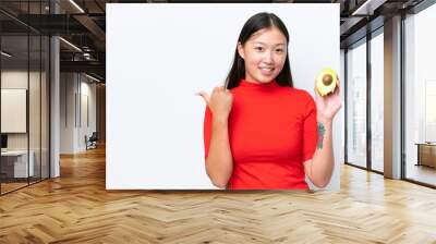 Young Asian woman holding an avocado isolated on white background pointing to the side to present a product Wall mural