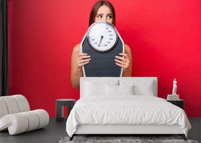 Young beautiful caucasian woman isolated on red background with weighing machine and hiding behind it Wall mural