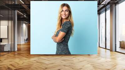 Young blonde Uruguayan isolated on blue background with arms crossed and happy Wall mural