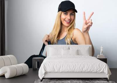 Young blonde woman holding a sport bag over isolated white background smiling and showing victory sign Wall mural