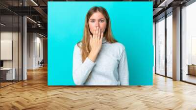 Young blonde woman isolated on blue background covering mouth with hand Wall mural