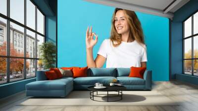 Young blonde woman isolated on blue background showing ok sign with fingers Wall mural