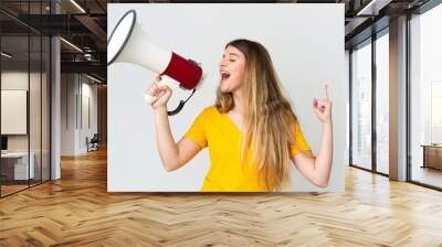 Young blonde woman isolated on white background shouting through a megaphone to announce something in lateral position Wall mural