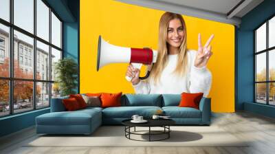 Young blonde woman isolated on yellow background holding a megaphone and smiling and showing victory sign Wall mural