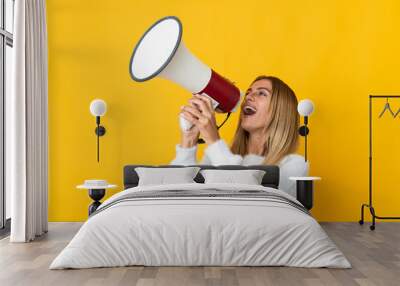 Young blonde woman isolated on yellow background shouting through a megaphone to announce something in lateral position Wall mural