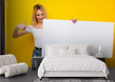 Young blonde woman over isolated yellow wall holding an empty white placard for insert a concept Wall mural